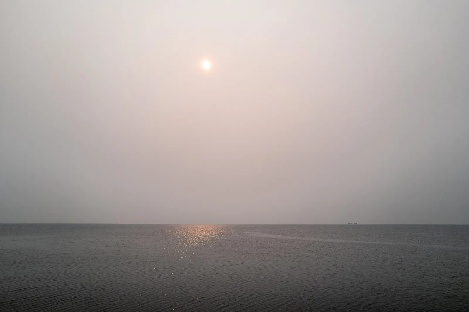A small pale sun in a hazy grey sky above a dark, flat lake, with a faint streak of light on the water